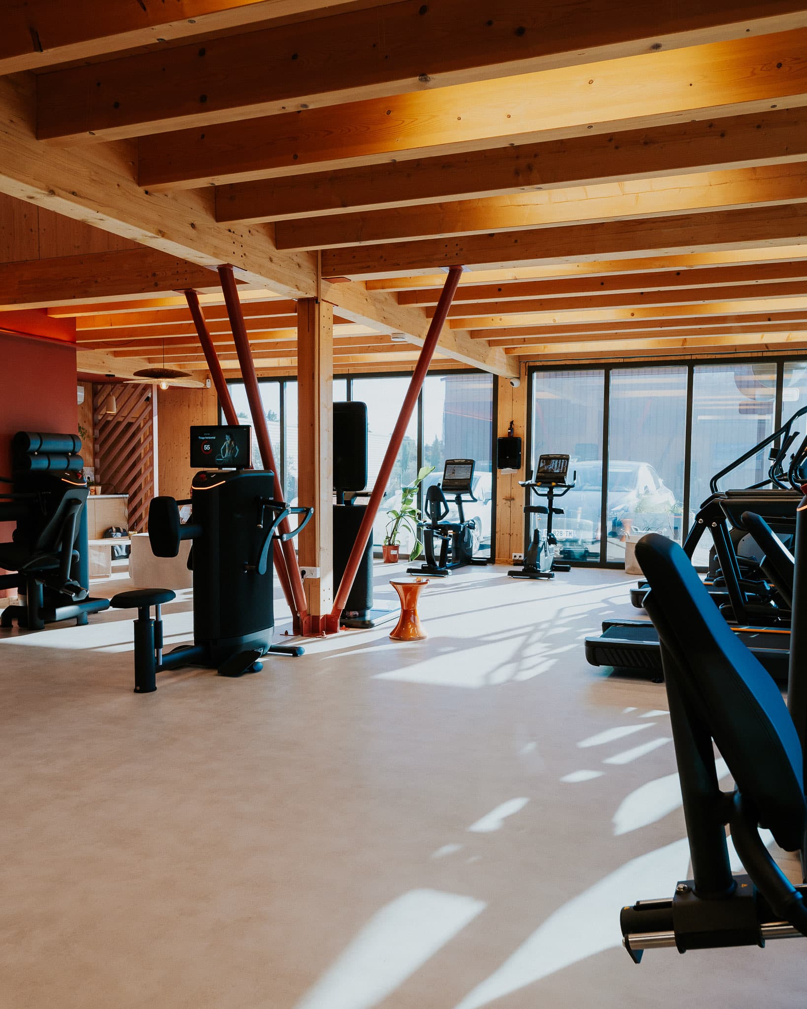 Plateau baigné de lumière : machines EGYM, poutres en bois et poteaux terracotta